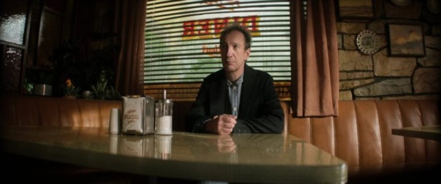 A man sitting alone at a diner booth with a neon "OPEN" sign in the background.