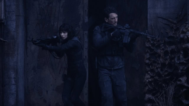 A woman and man aiming guns in a dark, forested setting for an action-packed scene.
