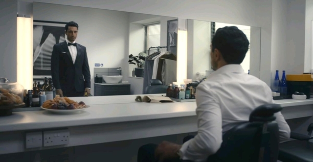 A man in a tuxedo looking at himself in a mirror in a dressing room. Riz Ahmed in Bait