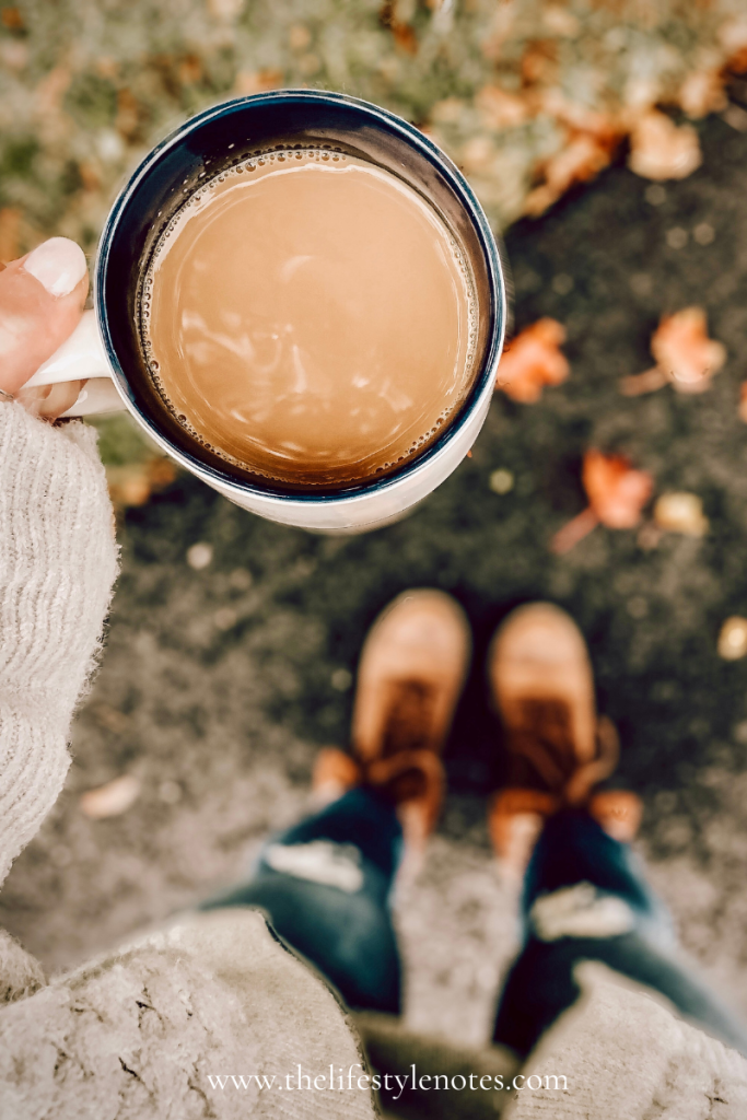 Fall Wardrobe Basics You'll Reach For All Season. Person holding a mug of coffee outdoors, with fall leaves on the ground.