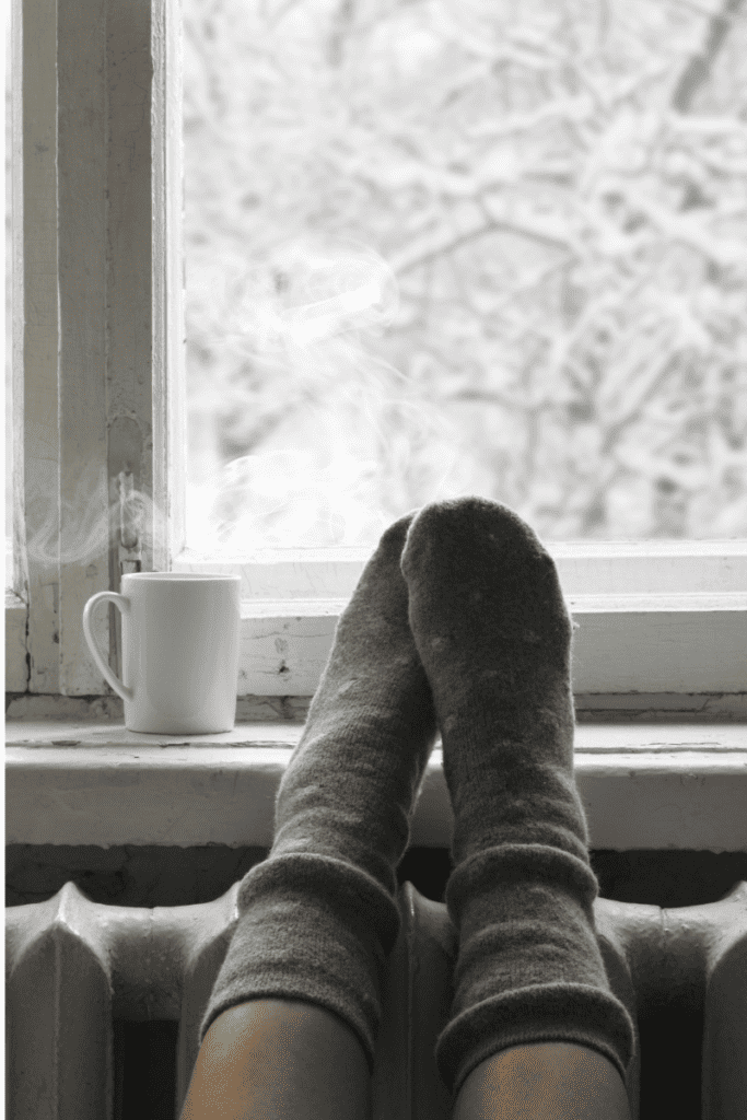 How To Take Care of Your Skin in the Winter. Feet up on a windowsill with snow outside. Cup of steaming coffee.