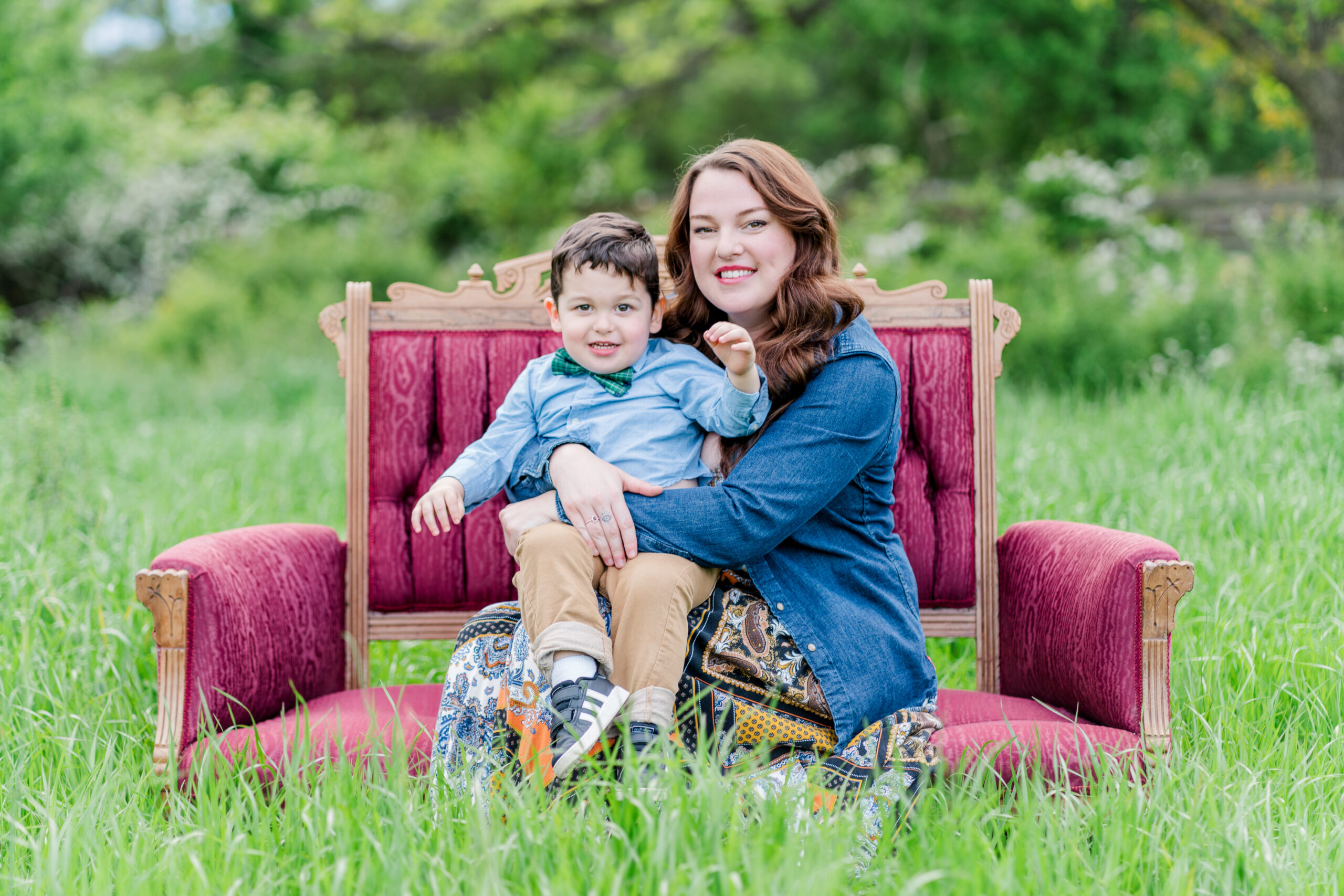 Mom Katy sits on a red vintage loveseat in a green field filled with tall grass. Her son Davis sits in her lap