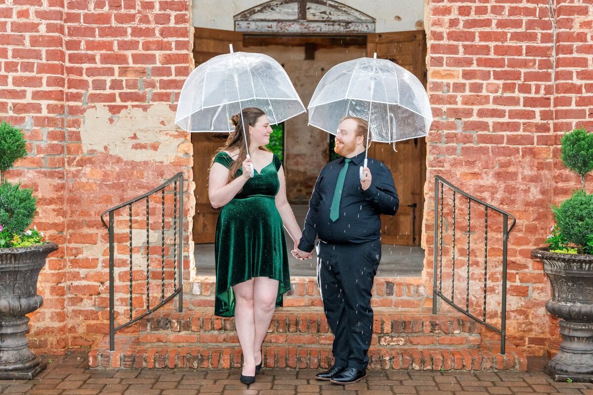 Couple laughing under clear umbrellas in the rain at a Providence Cotton Mill engagement session in Maiden NC