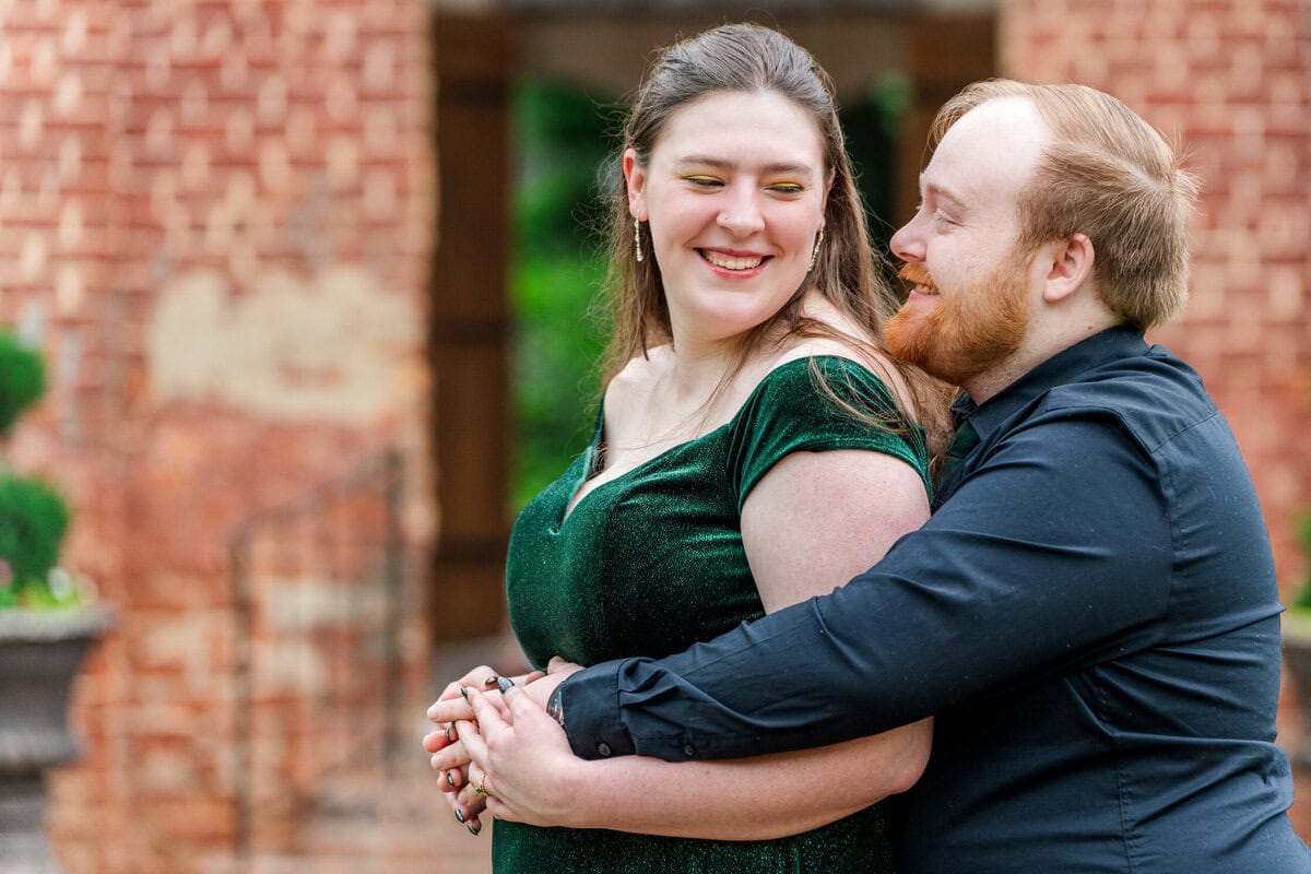 Bride-to-be and groom-to-be embracing inside historic cotton mill with exposed brick and arched windows