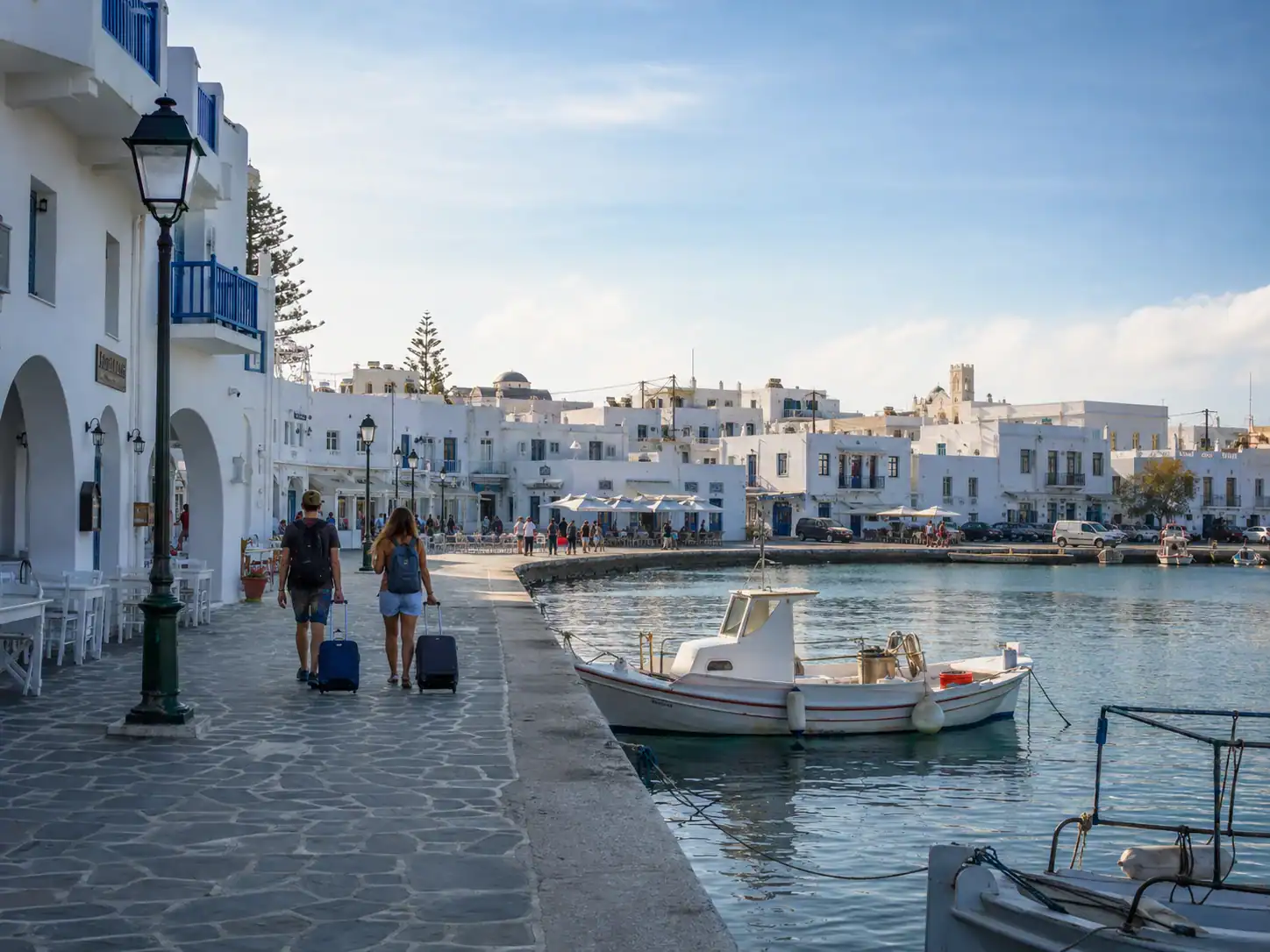 Cycladic harbor promenade with whitewashed buildings, fishing boats and travelers walking with luggage