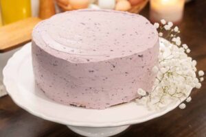 A phot of a finished blueberry cake with a fresh sprig of babys breath on a white pedestal cake stand in a cottage kitchen on a wooden counter top.