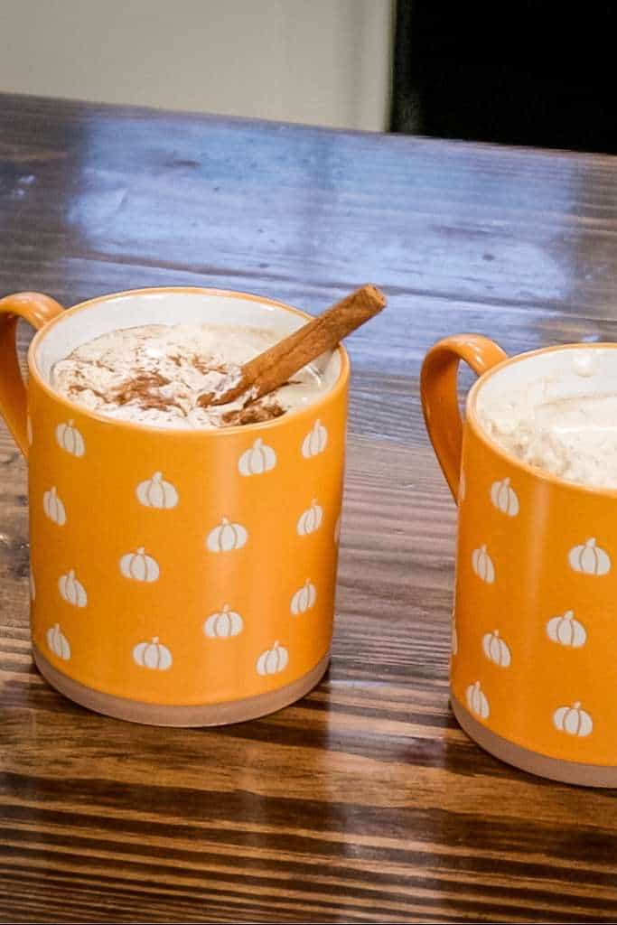 two orange coffee cups filled with pumpkin spice latte and a cinnamon stick on a wooden counter top.