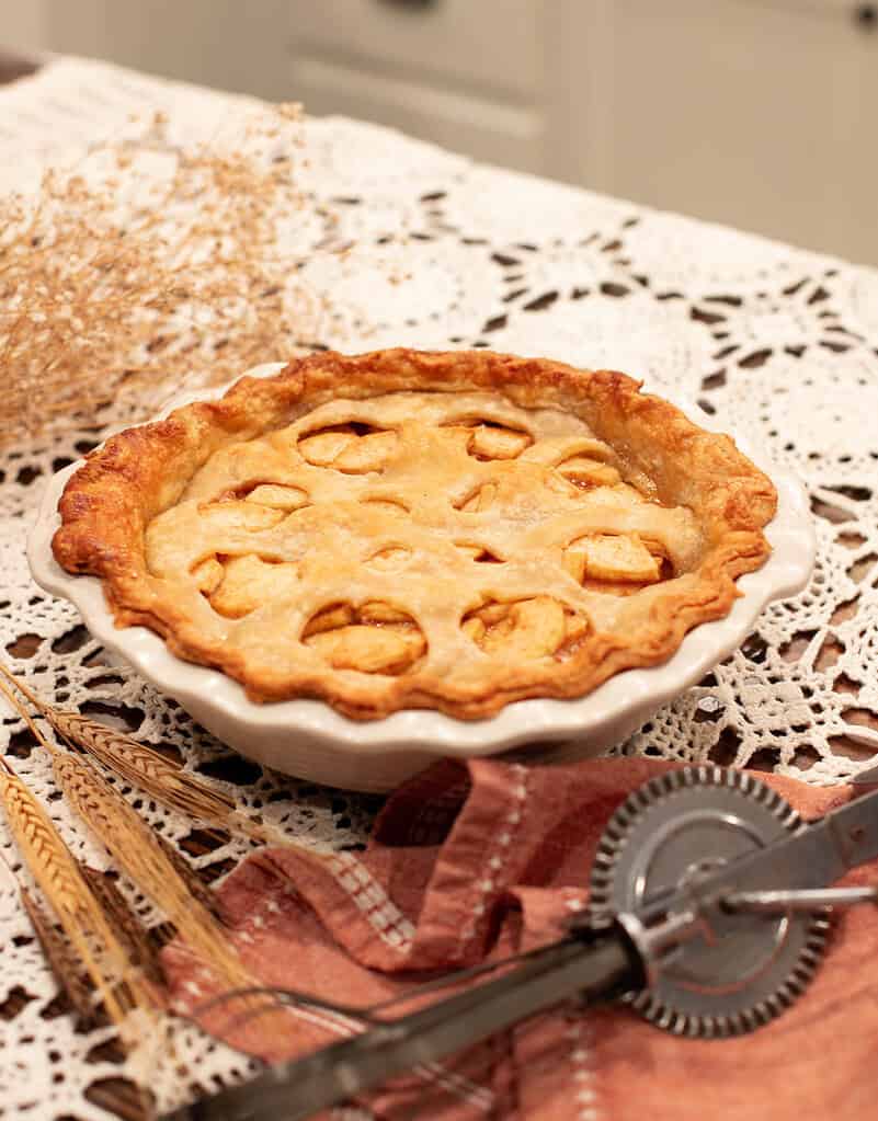 Baked sourdough apple pie with a golden crust and small cutouts on top, in a white pie dish, with wheat stalks and vintage baking tools nearby on a lace tablecloth.