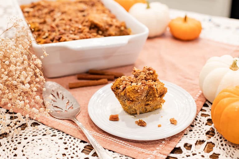 A slice of sourdough pumpkin French toast casserole on a plate with cinnamon sticks and a casserole dish in the background, accompanied by autumn decor.