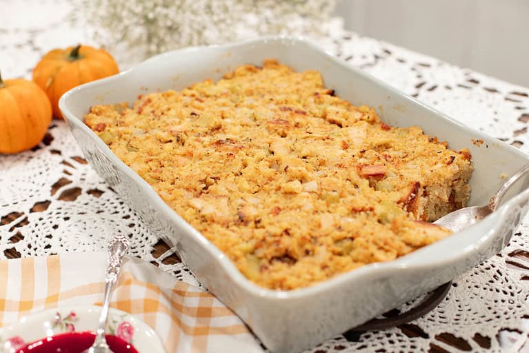 A close-up of the baked sourdough cornbread dressing in a ceramic baking dish, with a spoon ready to serve. The dish’s golden-brown texture captures the rustic appeal of a comforting holiday side dish.