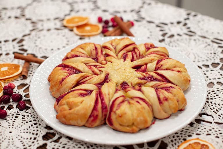A side view of the cranberry orange star bread, showing the soft, fluffy texture and vibrant cranberry filling. The table setting includes crocheted lace and festive garnishes like dried oranges and cranberries.
