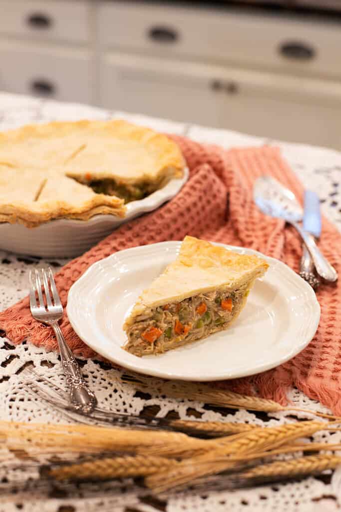A cozy table setting featuring a slice of chicken pot pie on a white plate, with the rest of the pie in the background, set on a lace tablecloth with a textured pink napkin.