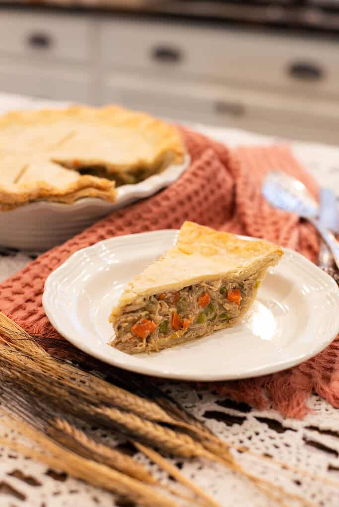 A slice of chicken pot pie on a white plate, highlighting the flaky crust and hearty filling, styled with wheat stalks and vintage silverware for a rustic touch.