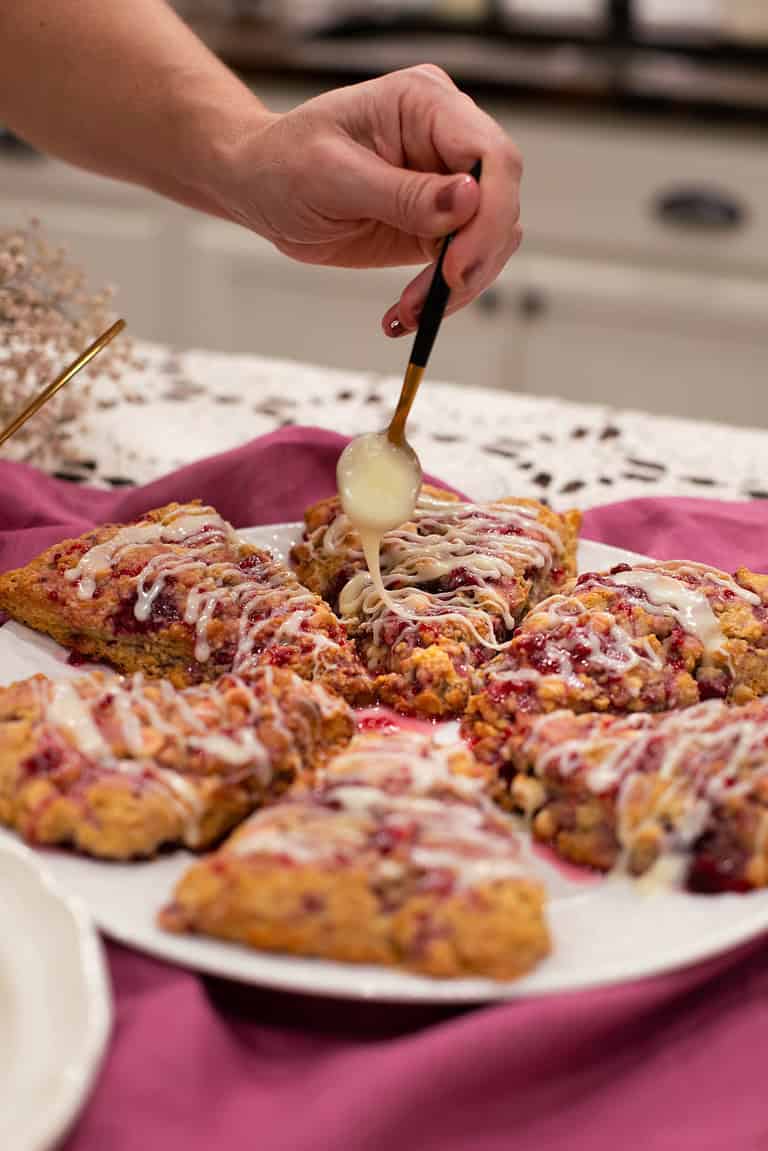 A hand lifting a glazed raspberry white chocolate sourdough scone from a plate, highlighting its texture and layers.