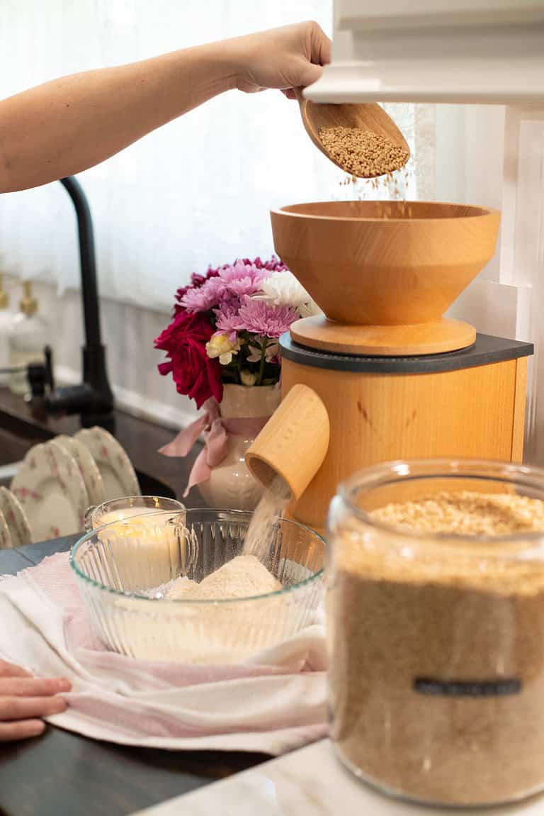 A hand pours hard white wheat berries into the wooden hopper of a grain mill as freshly milled flour falls into a glass bowl below. A vase of pink and red flowers sits nearby, creating a cozy kitchen atmosphere.