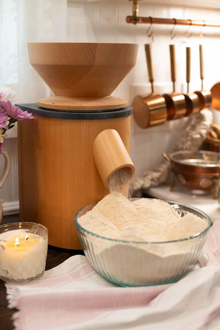 Close-up of a grain mill actively grinding flour into a large bowl. The bowl is now filled with fluffy mounds of flour. Warm kitchen tones and copper utensils accent the scene.