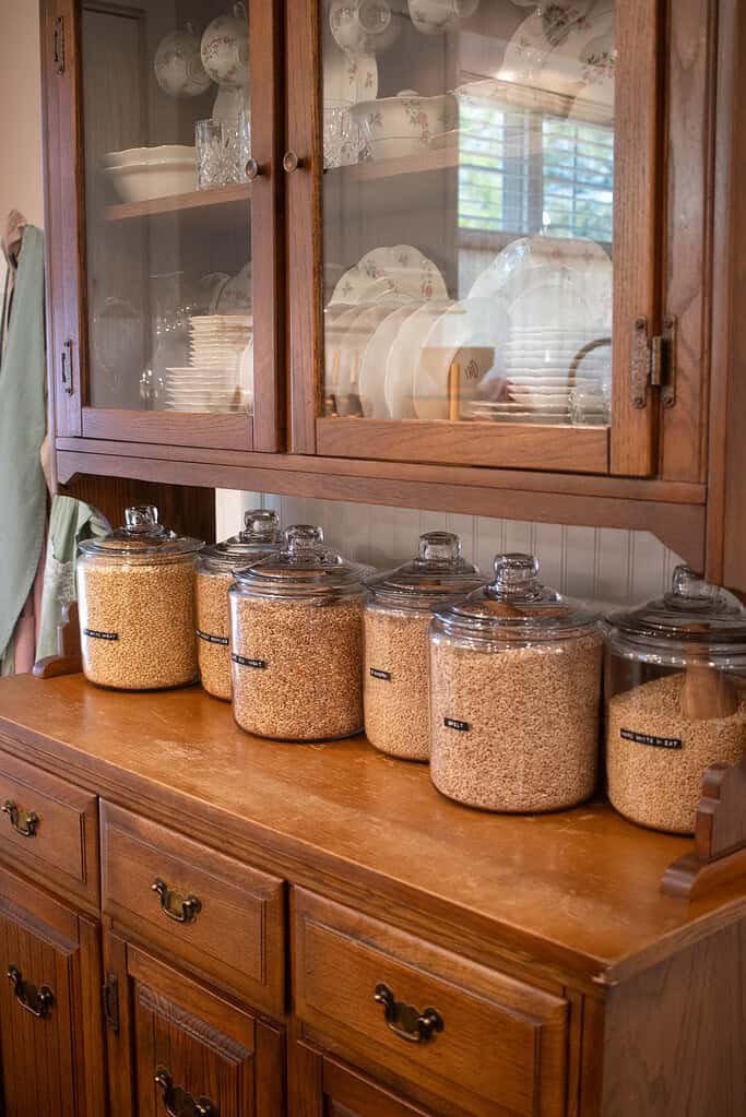 Five large glass jars filled with various wheat berries, all labeled (e.g., “EINKORN,” “HARD WHITE,” “SPELT”), sit lined up on a wooden sideboard beneath a china cabinet filled with floral porcelain dishes.
