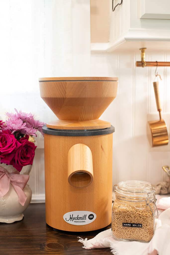 A wooden Mockmill 200 grain mill sits on a dark counter with a clear glass jar labeled “HARD WHITE” beside it. The mill is flanked by a bouquet of roses and copper kitchenware in the background.