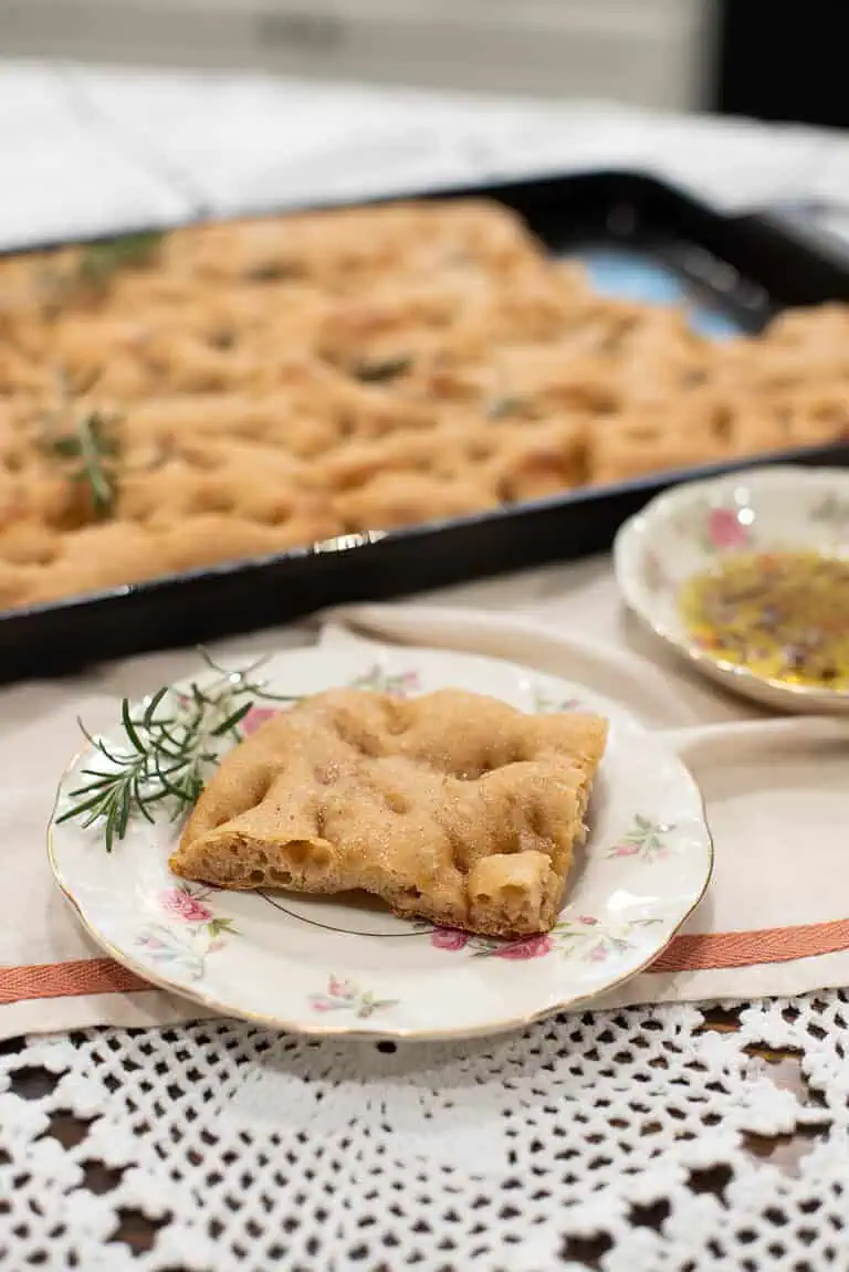 A single slice of golden-brown sourdough focaccia sits on a vintage floral plate, garnished with fresh rosemary. The full tray of focaccia is in the background, with a dish of herb-infused olive oil for dipping.