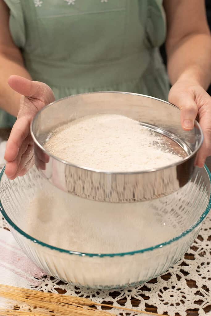 Person in a green dress manually sifting freshly milled flour over a glass bowl using a round stainless steel sieve.