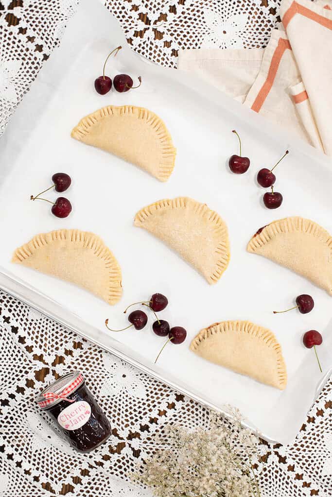 A baking sheet lined with parchment paper, holding unbaked cherry hand pies with crimped edges, surrounded by fresh cherries and a jar of cherry jam.