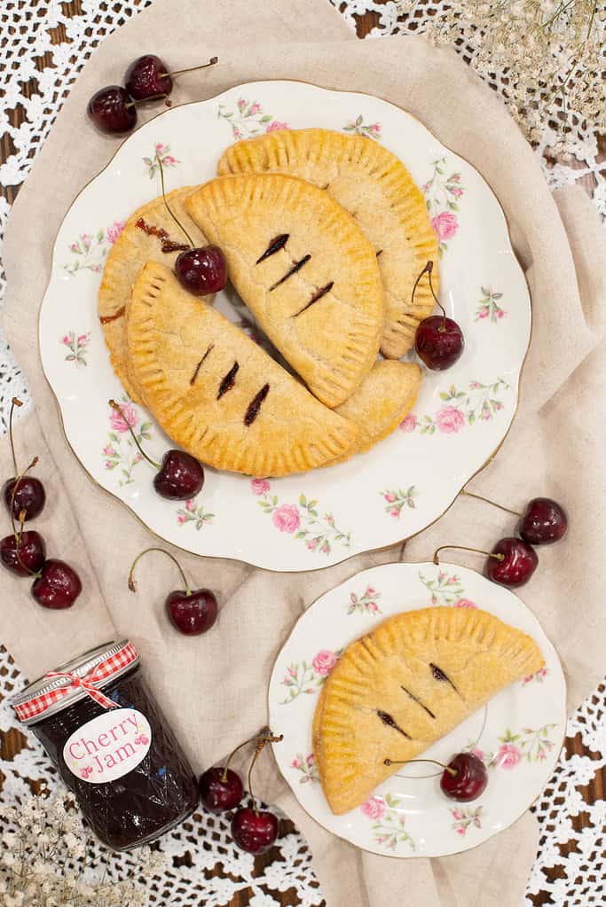 A floral plate stacked with golden, freshly baked cherry hand pies, with a jar of cherry jam and fresh cherries beside it.