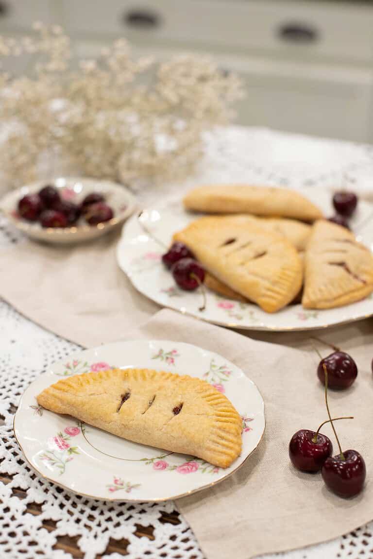 A cozy kitchen scene featuring a plate of cherry hand pies, fresh cherries, and delicate dried flowers.