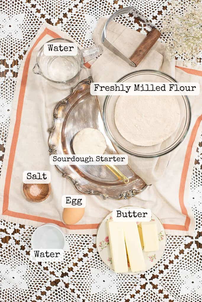 A flat lay of sourdough pastry ingredients, including freshly milled flour, sourdough starter, butter, egg, salt, and water, arranged on a linen cloth with vintage kitchen tools. Labels identify each ingredient.