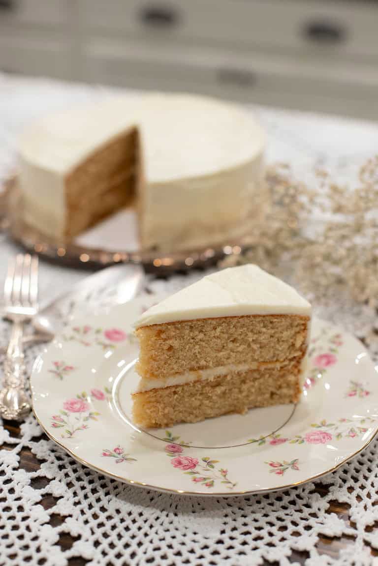 A beautifully plated slice of vanilla cake with buttercream frosting, with a blurred background of the remaining cake and delicate baby’s breath flowers.