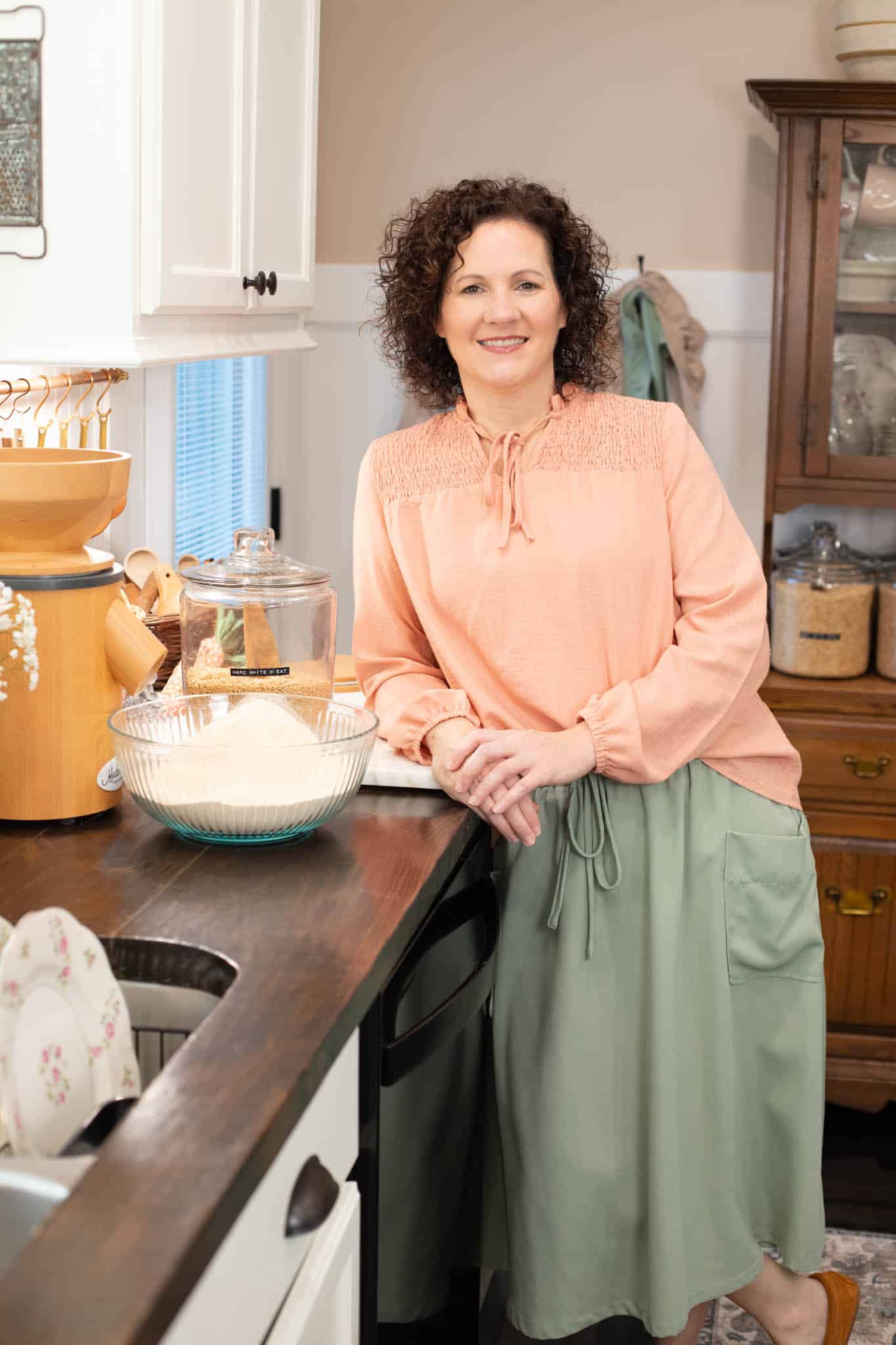 Close-up of a woman smiling in a peach blouse and sage green skirt, standing near a jar of hard white wheat and a bowl of flour.