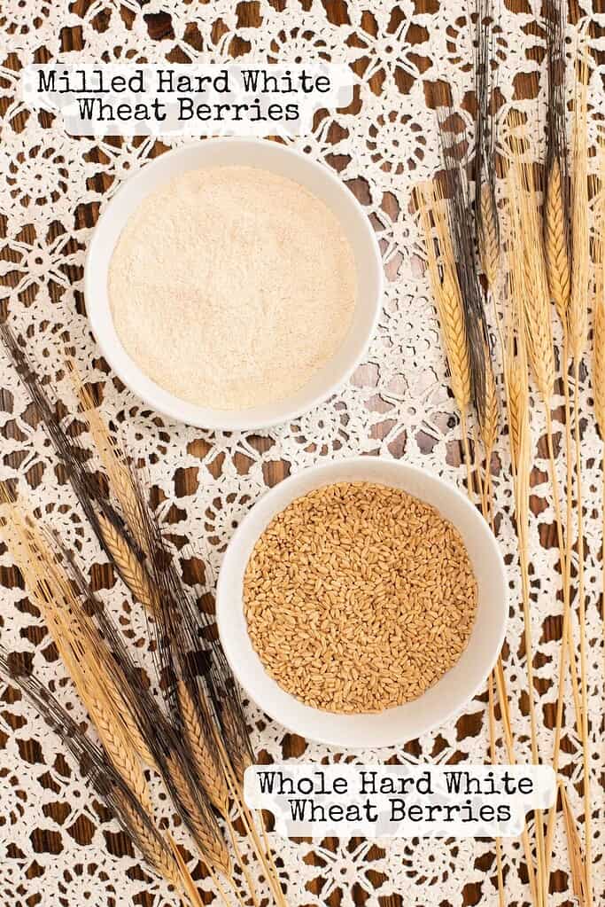 Top-down view of milled hard white wheat berries and whole hard white wheat berries in white bowls on a lace tablecloth, surrounded by wheat stalks.