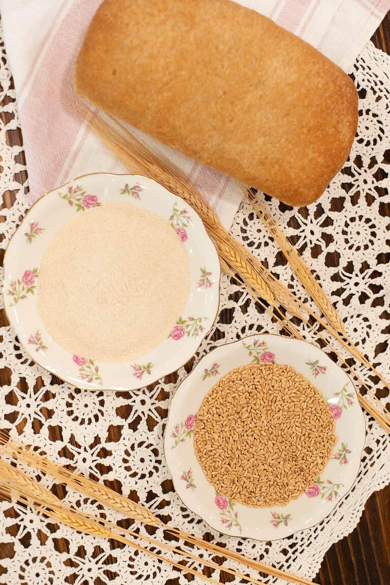 Overhead view of a loaf of freshly baked whole wheat bread on a pink-striped cloth napkin. Below it are two floral china plates—one filled with freshly milled whole wheat flour, and the other with whole wheat berries—set atop a vintage lace tablecloth with decorative wheat stalks arranged nearby.