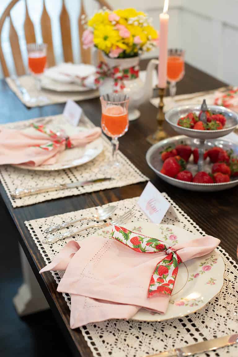 Angled view of a place setting featuring a pink napkin tied with strawberry-printed ribbon on a floral plate atop a lace placemat.