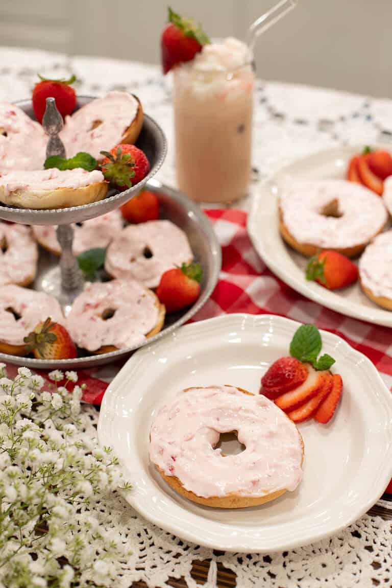 Bagels generously topped with whipped strawberry cream cheese served on white plates and a tiered tray, garnished with fresh strawberries and mint leaves.