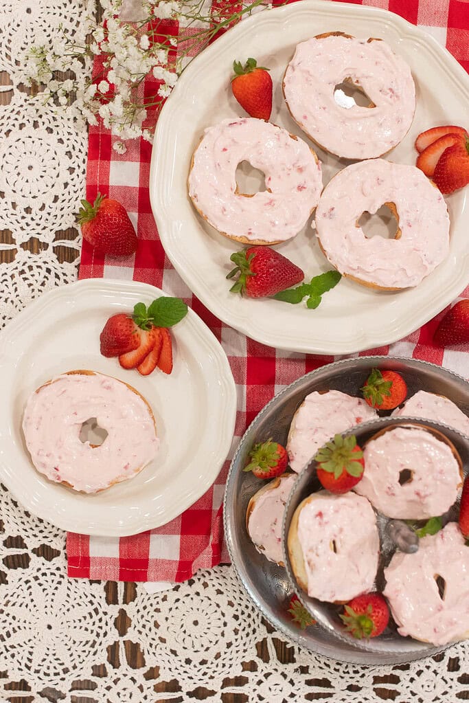 Overhead view of several bagels topped with strawberry cream cheese on white plates and a silver tiered stand, surrounded by fresh strawberries and baby’s breath flowers.