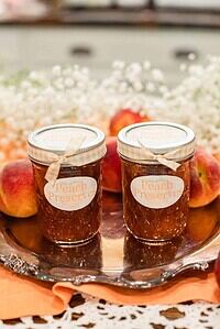 Two mason jars labeled “Peach Preserves” with gingham ribbons, surrounded by fresh peaches and baby’s breath flowers on a silver tray.