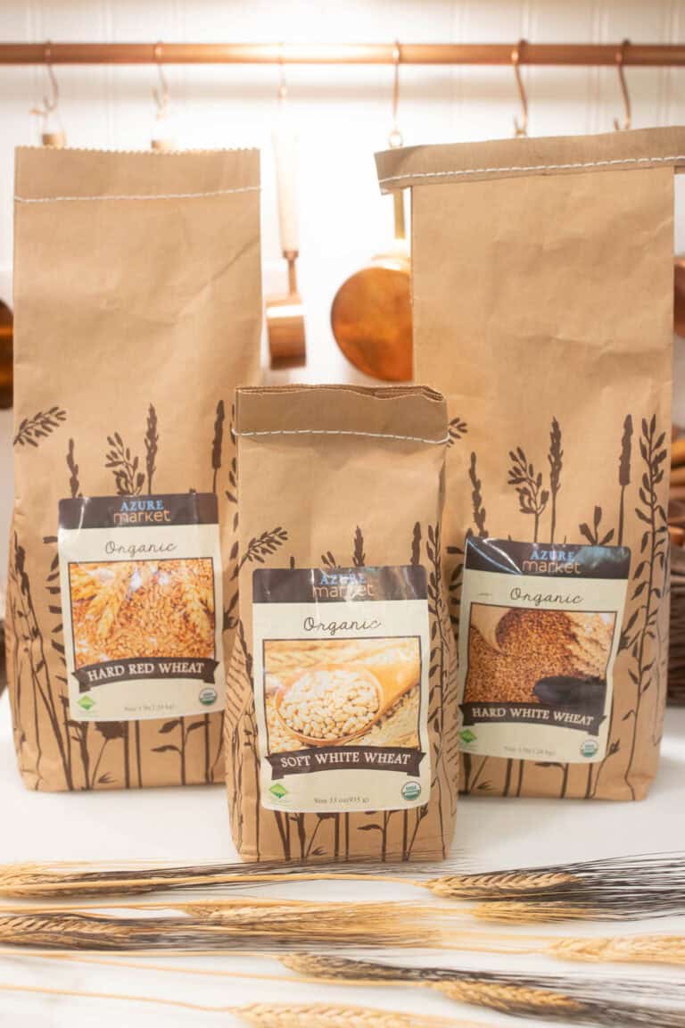 Three kraft paper bags of organic wheat berries from Azure Standard, including hard red wheat, hard white wheat, and soft white wheat, styled on a white counter with vintage copper pots and wheat stalks.