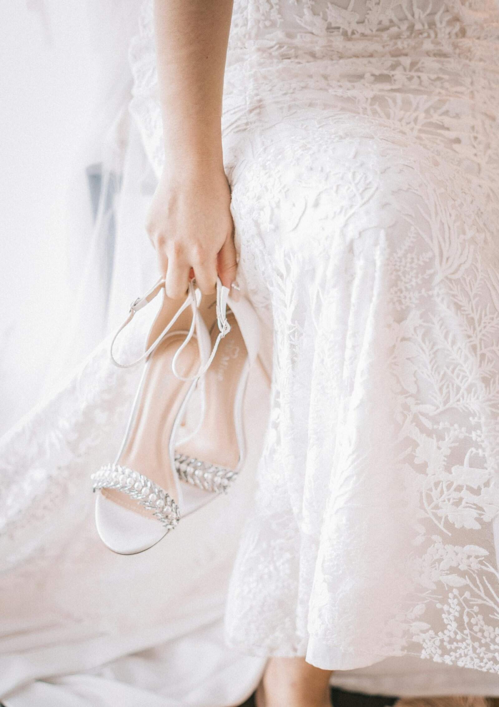 Close-up of woman holding bridal shoes wearing elegant white dress.