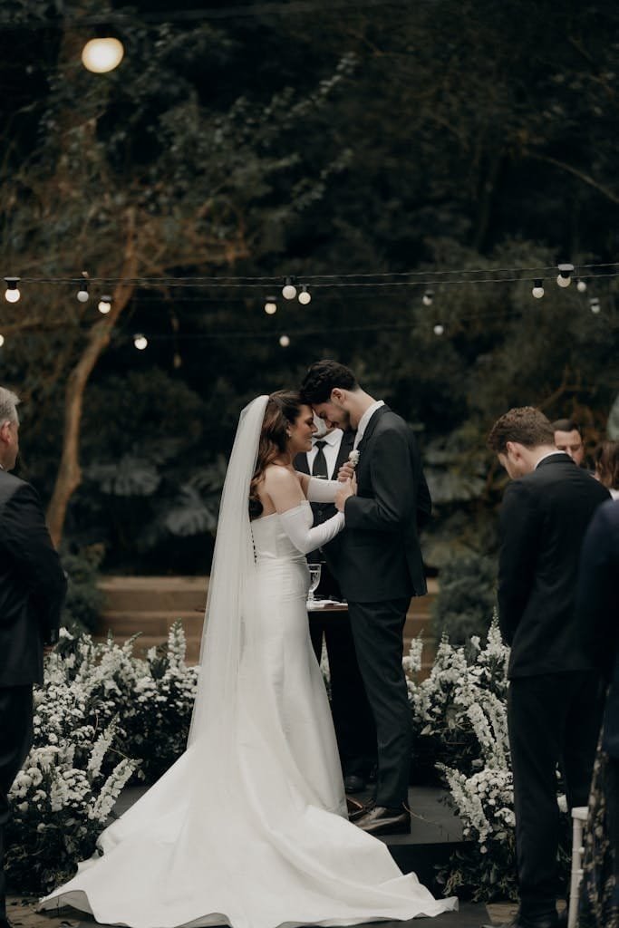 A romantic outdoor wedding ceremony with a bride and groom exchanging vows under string lights at twilight.
