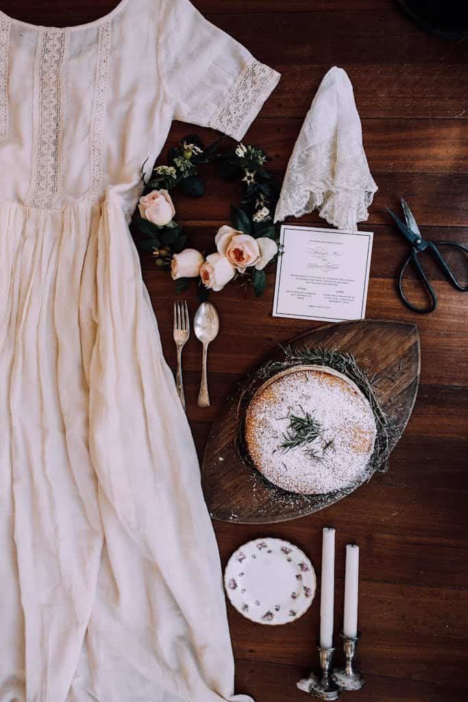 An elegant flat lay featuring a lace wedding dress, cake, and floral decor.