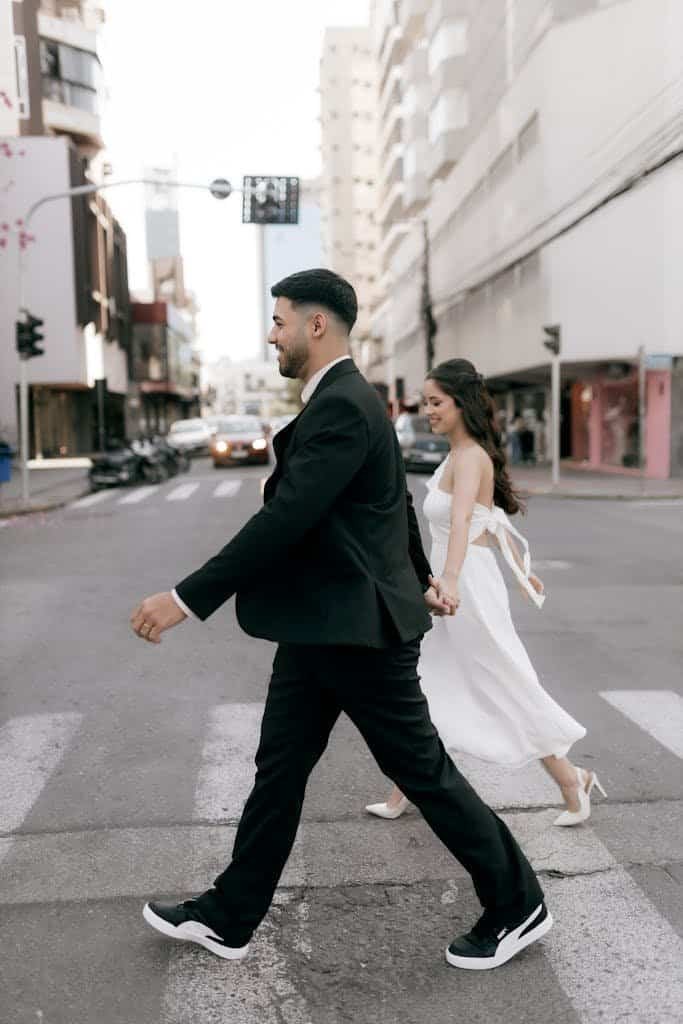Bride and groom crossing city street in wedding attire, holding hands.