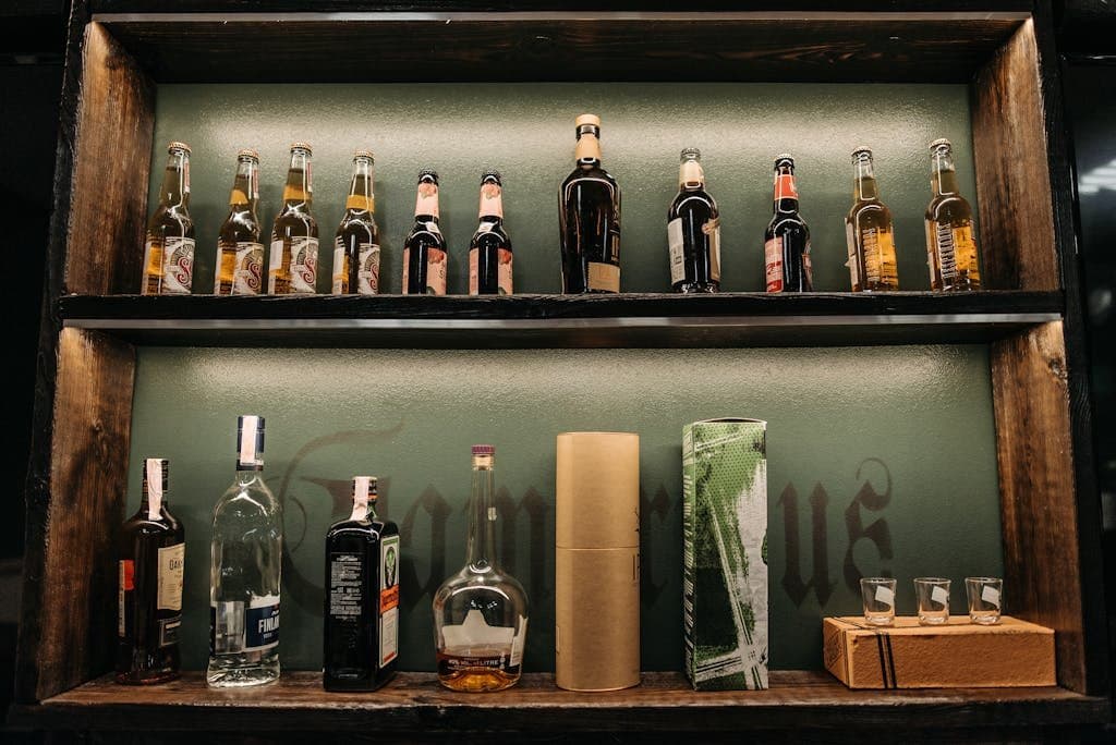 Rustic bar shelves displaying a variety of alcohol bottles, including beer and spirits, with shot glasses.