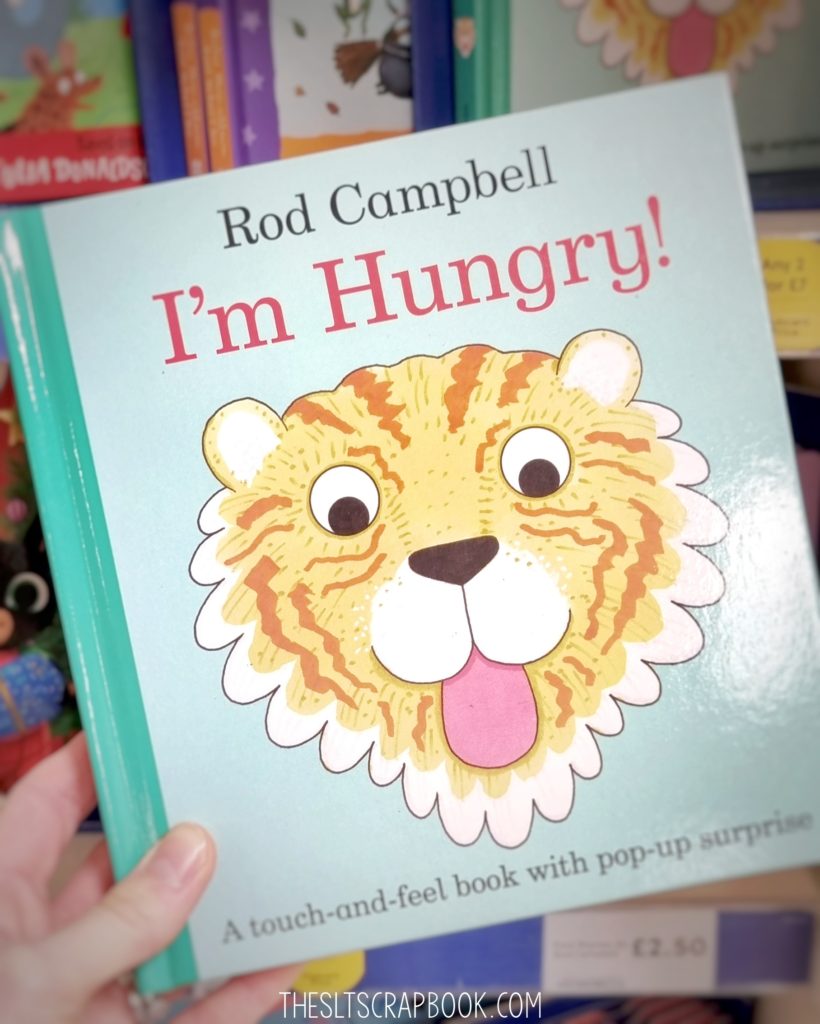 Picture of the 'I'm Hungry' food-themed book by Rod Campbell in the foreground. The background is blurred with a range of other books shown on the shelves.