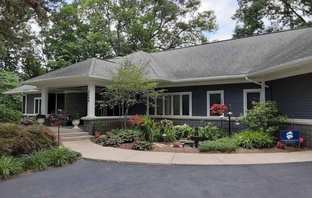 Elegant home exterior with professionally painted navy blue siding, lush landscaping, and decorative elements enhancing curb appeal | Thornapple Painting logo visible in garden area.
