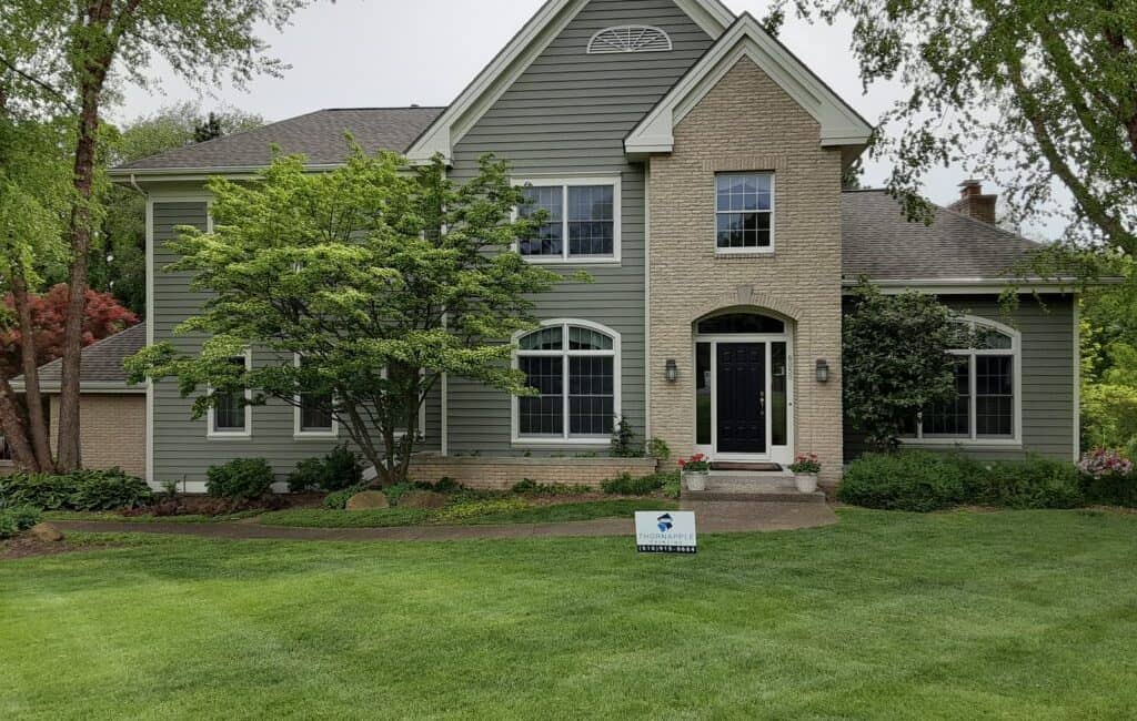 Spacious residential house with modern green siding and brick accents, surrounded by lush landscaping and mature trees, showcasing professional exterior painting services.