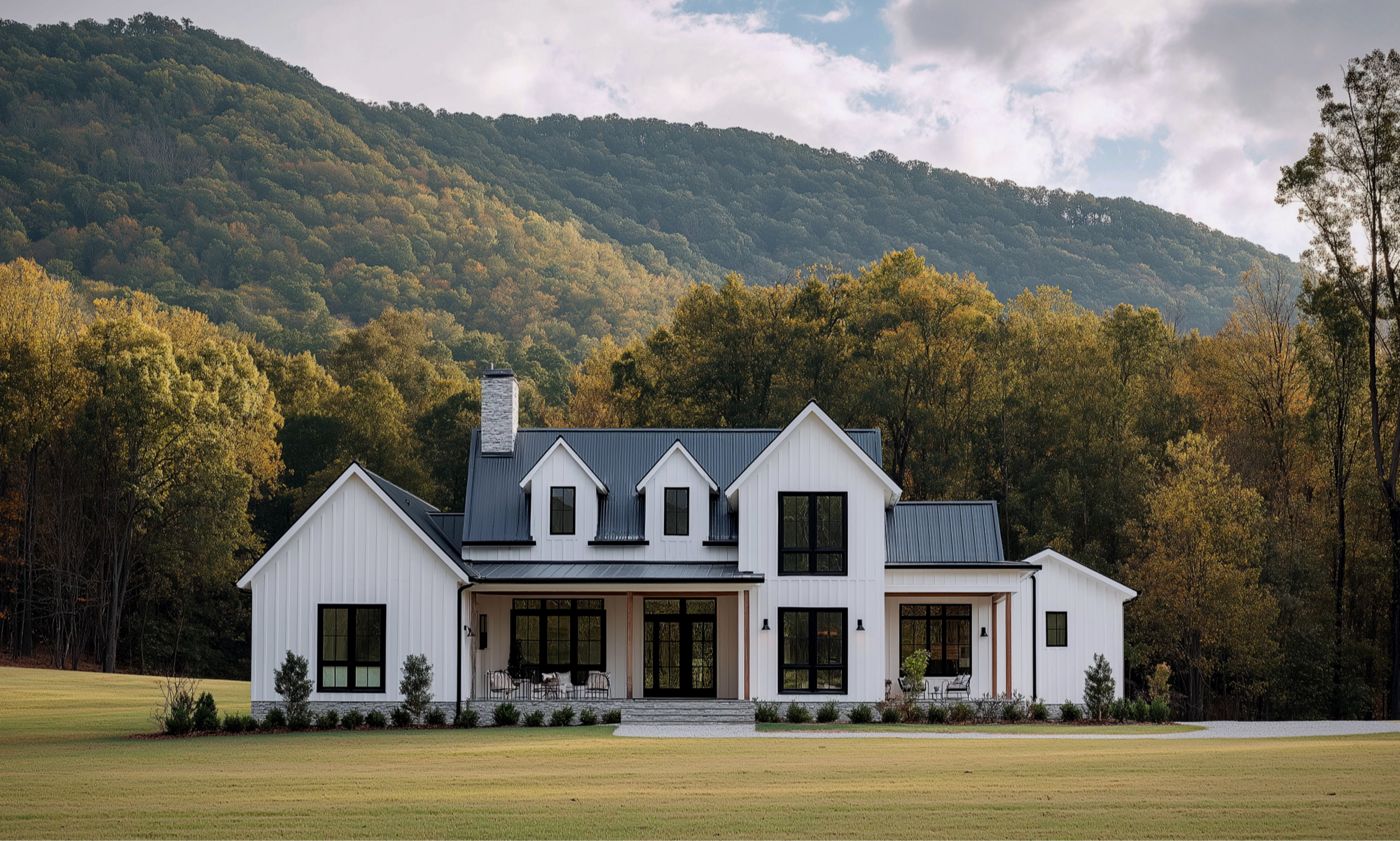 White modern farmhouse with black roof against a forested mountain.