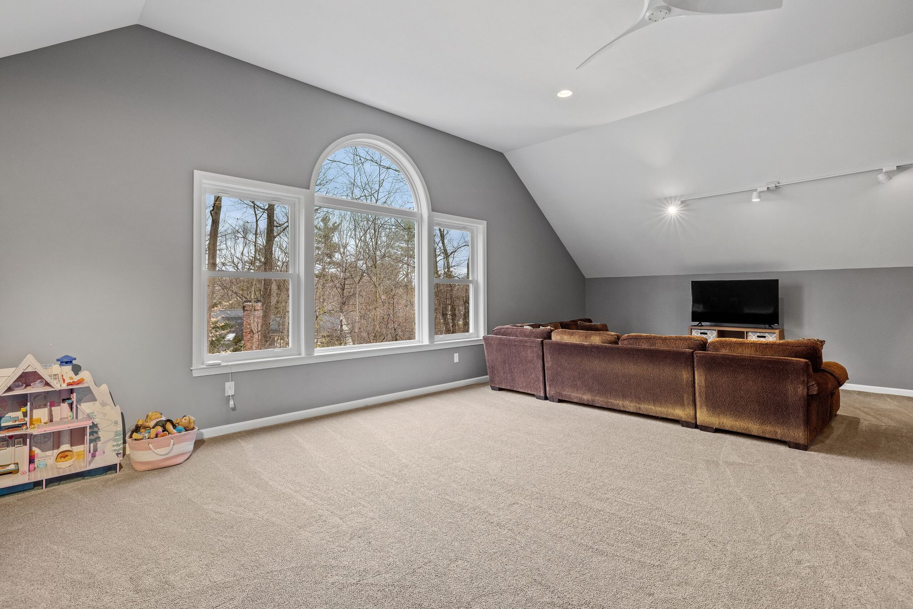Spacious attic room with large arched window, brown sectional sofa, and children's toys.
