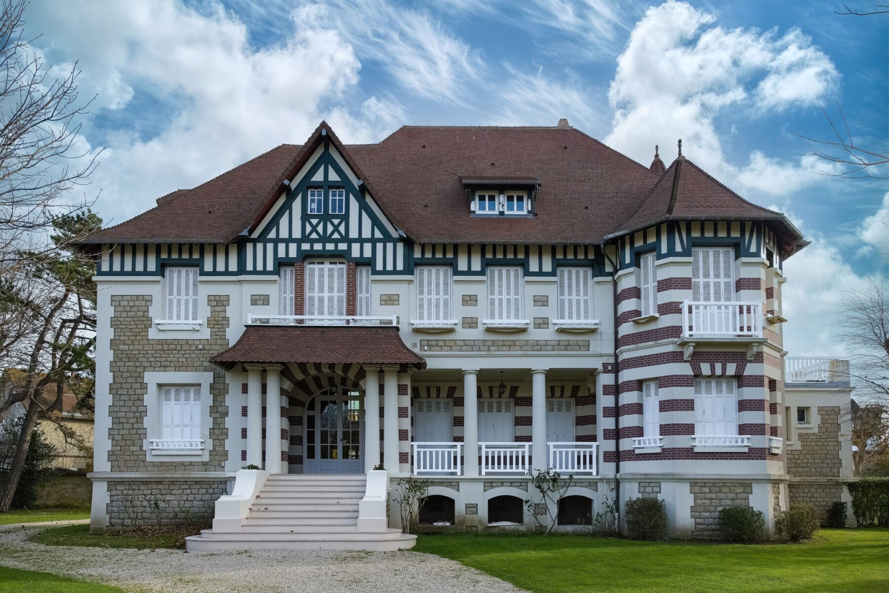 Large, ornate house with a red roof and white shutters against a cloudy sky.