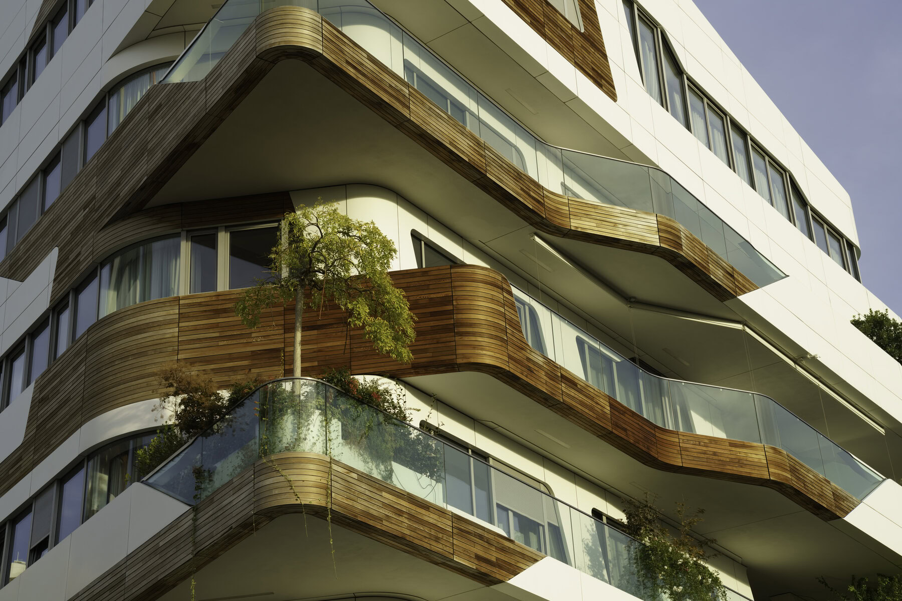 Modern building with curved balconies, glass railings, and wooden accents, featuring small trees.