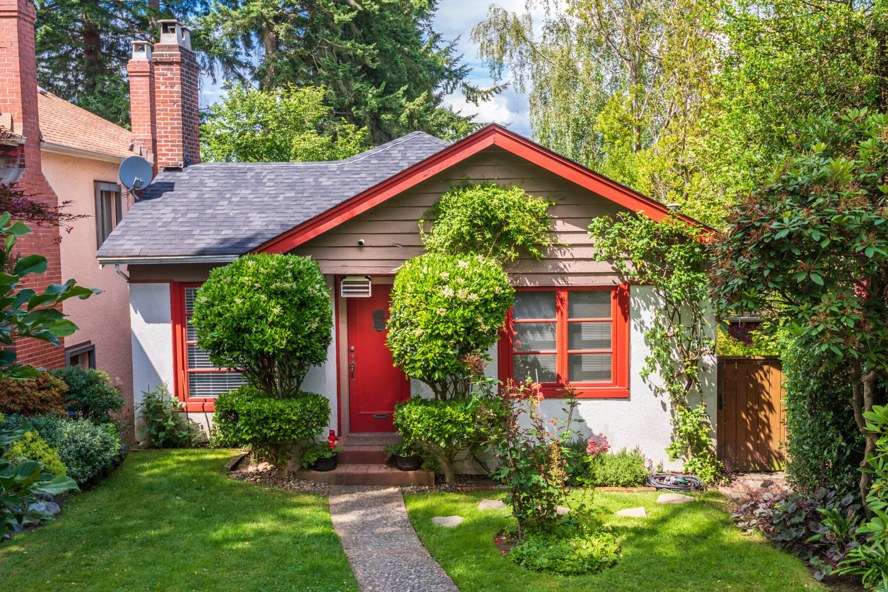 Small house with red door and windows, surrounded by green trees and bushes.